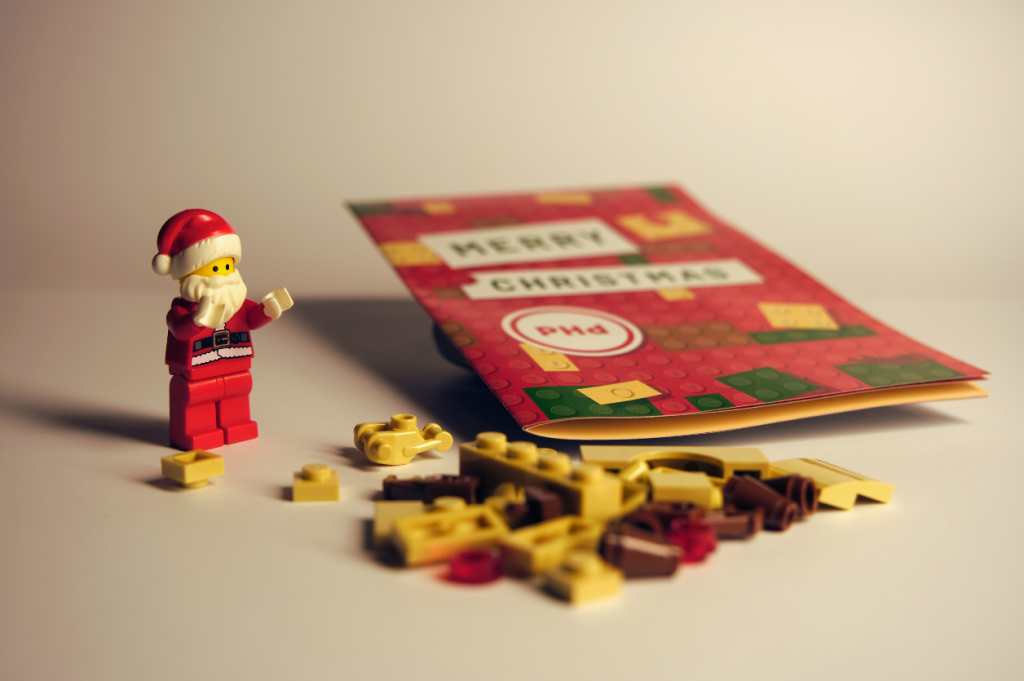 LEGO Santa figure standing beside scattered Rudolph pieces in front of a PHd Design Christmas leaflet, promoting the festive LEGO advent build.